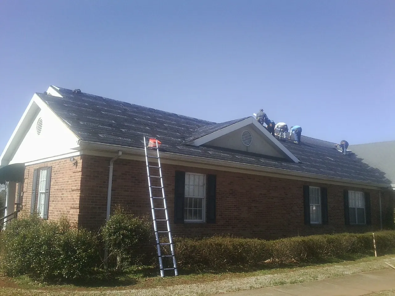 Crew tearing shingles off a brick ranch roof with a ladder set — Emergency Roof Repair work by our crew in Aledo